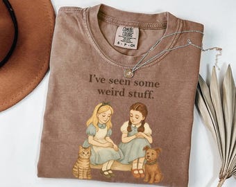 May include: Brown t-shirt with the text "I've seen some weird stuff." The shirt features an illustration of two girls, a cat, and a dog. A silver necklace with a pendant is draped across the shirt.