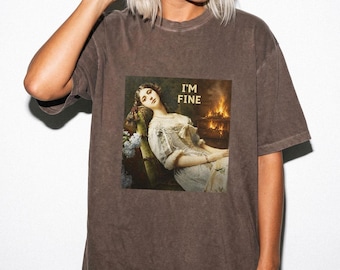 May include: A brown short-sleeve t-shirt featuring a graphic print of a painting showing a person resting in a chair near a fireplace with the text I'M FINE printed in gold letters above the scene. The shirt has a crew neckline and a relaxed fit.