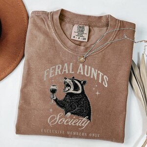 May include: A brown Comfort Colors t-shirt with the words "FERAL AUNTS Society" and a raccoon graphic holding a wine glass. The shirt also has the text "EXCLUSIVE MEMBERS ONLY". A silver necklace with a pendant is on the shirt.