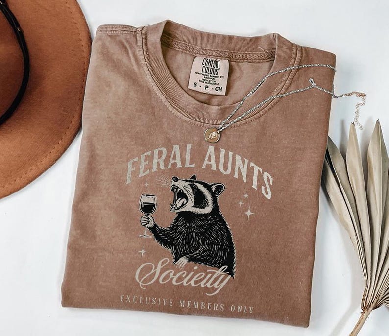 May include: A brown Comfort Colors t-shirt with the words "FERAL AUNTS Society" and a raccoon graphic holding a wine glass. The shirt also has the text "EXCLUSIVE MEMBERS ONLY". A silver necklace with a pendant is on the shirt.