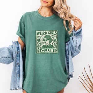 May include: Green t-shirt with the text "WEIRD GIRLS CLUB" in a square design. The design features a woman wearing sunglasses and holding a cup. The shirt is paired with a denim jacket.