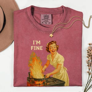 May include: A dusty rose Comfort Colors t-shirt with the phrase "I'M FINE" and a vintage illustration of a woman cooking with flames. A gold star pendant necklace and a brown hat are also in the image.