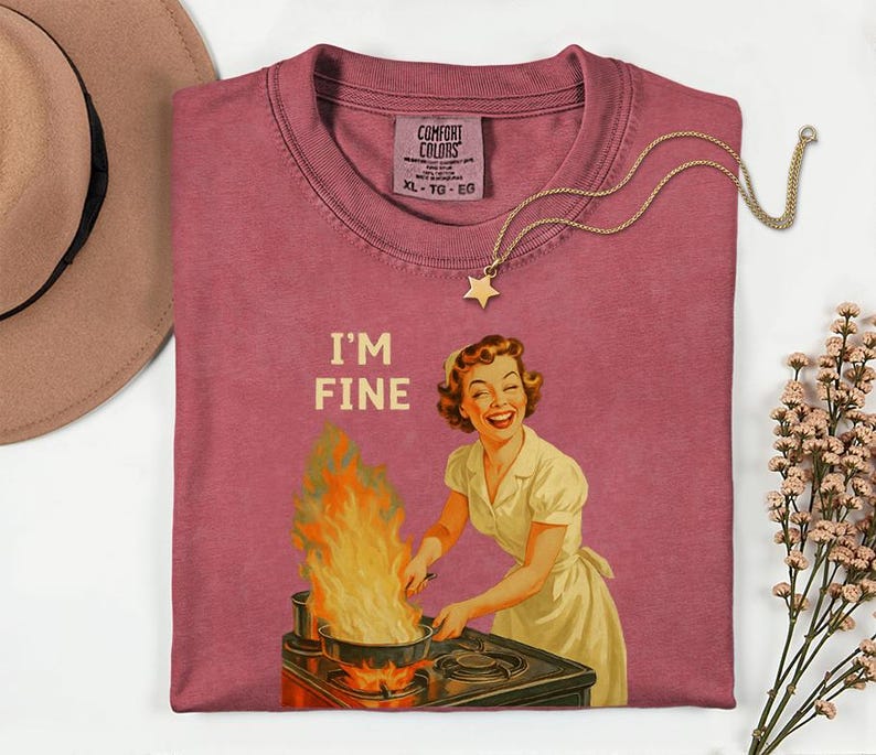 May include: A dusty rose Comfort Colors t-shirt with the phrase "I'M FINE" and a vintage illustration of a woman cooking with flames. A gold star pendant necklace and a brown hat are also in the image.