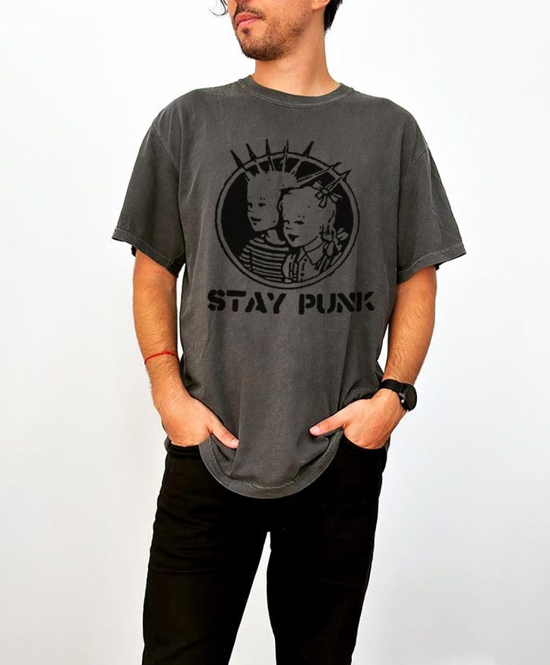 May include: A gray t-shirt with a black graphic of two children with a mohawk and a bow. The graphic is inside a circle with the words "STAY PUNK" below it.