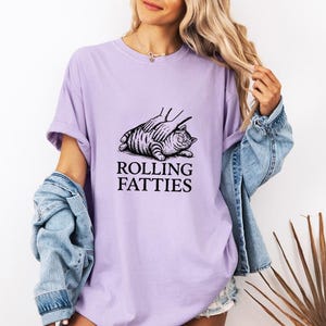 May include: A light purple t-shirt with the text "ROLLING FATTIES" below a black and white illustration of a cat. The cat is lying down with a hand on its back. The person is wearing a denim jacket.