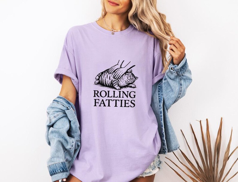 May include: A light purple t-shirt with the text "ROLLING FATTIES" below a black and white illustration of a cat. The cat is lying down with a hand on its back. The person is wearing a denim jacket.