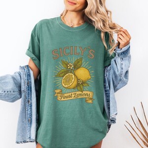May include: Green t-shirt with a vintage-style graphic featuring lemons, leaves, and the text "Sicily's Finest Lemons." The design includes a whole lemon, a sliced lemon, and a banner. The shirt is worn with a denim jacket.