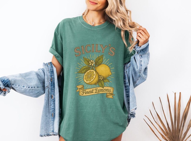 May include: Green t-shirt with a vintage-style graphic featuring lemons, leaves, and the text "Sicily's Finest Lemons." The design includes a whole lemon, a sliced lemon, and a banner. The shirt is worn with a denim jacket.