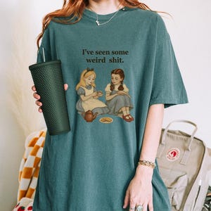 May include: Teal-colored t-shirt with the text "I've seen some weird shit." The graphic depicts two girls having tea. The person is holding a dark green textured tumbler.