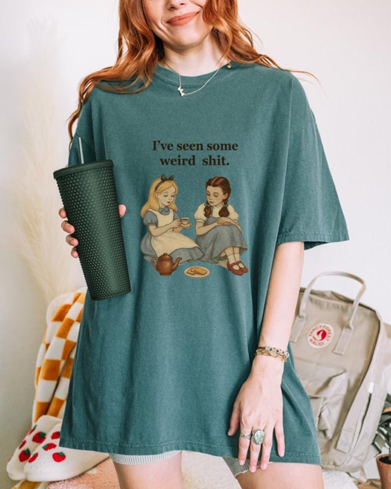 May include: Teal-colored t-shirt with the text "I've seen some weird shit." The graphic depicts two girls having tea. The person is holding a dark green textured tumbler.