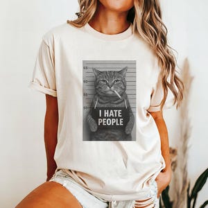 May include: Beige t-shirt featuring a black and white image of a cat holding a sign that reads "I HATE PEOPLE" with a cigarette in its mouth. The cat is positioned in front of a height chart.