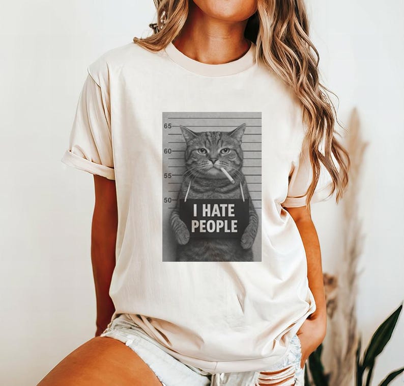 May include: Beige t-shirt featuring a black and white image of a cat holding a sign that reads "I HATE PEOPLE" with a cigarette in its mouth. The cat is positioned in front of a height chart.
