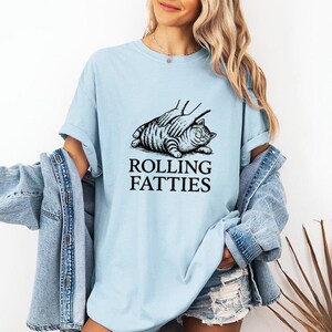 May include: Light blue t-shirt featuring a black and white illustration of a cat being petted, with the text "ROLLING FATTIES" below. The shirt has short sleeves and a relaxed fit.