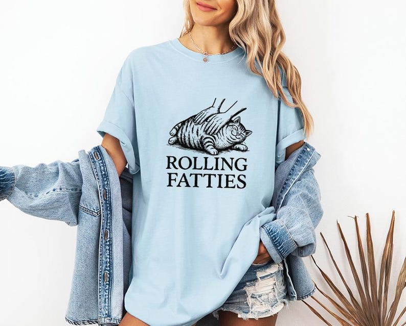 May include: Light blue t-shirt featuring a black and white illustration of a cat being petted, with the text "ROLLING FATTIES" below. The shirt has short sleeves and a relaxed fit.