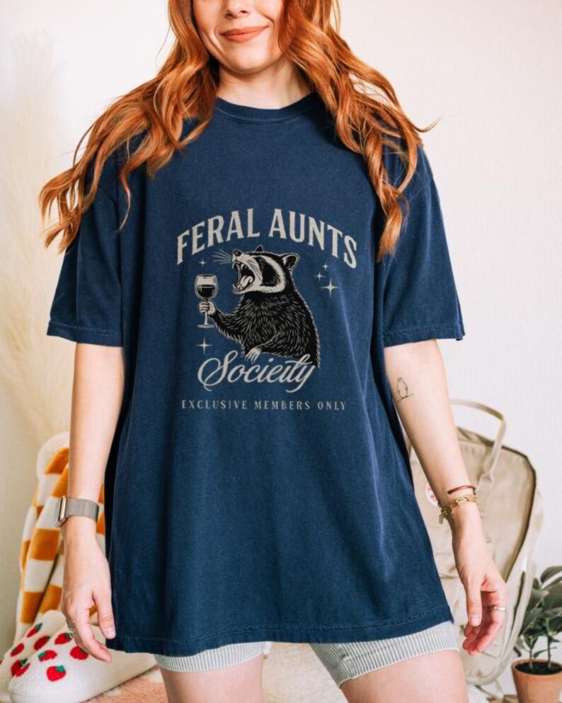 May include: Navy blue t-shirt with the text "FERAL AUNTS Society" and a graphic of a raccoon holding a wine glass. The shirt has short sleeves and a relaxed fit. The words "EXCLUSIVE MEMBERS ONLY" are printed below the word "Society".