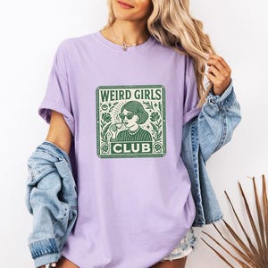 May include: A light purple t-shirt with a green graphic that reads "WEIRD GIRLS CLUB." The graphic features a woman wearing sunglasses and holding a cup. The shirt is made of a soft material.