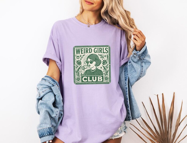 May include: A light purple t-shirt with a green graphic that reads "WEIRD GIRLS CLUB." The graphic features a woman wearing sunglasses and holding a cup. The shirt is made of a soft material.