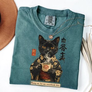 May include: Teal Comfort Colors t-shirt featuring a black cat in a kimono holding a coffee cup. The shirt has Japanese text and the phrase "Hang on, let me overthink this." A gold necklace and a hat are also in the image.