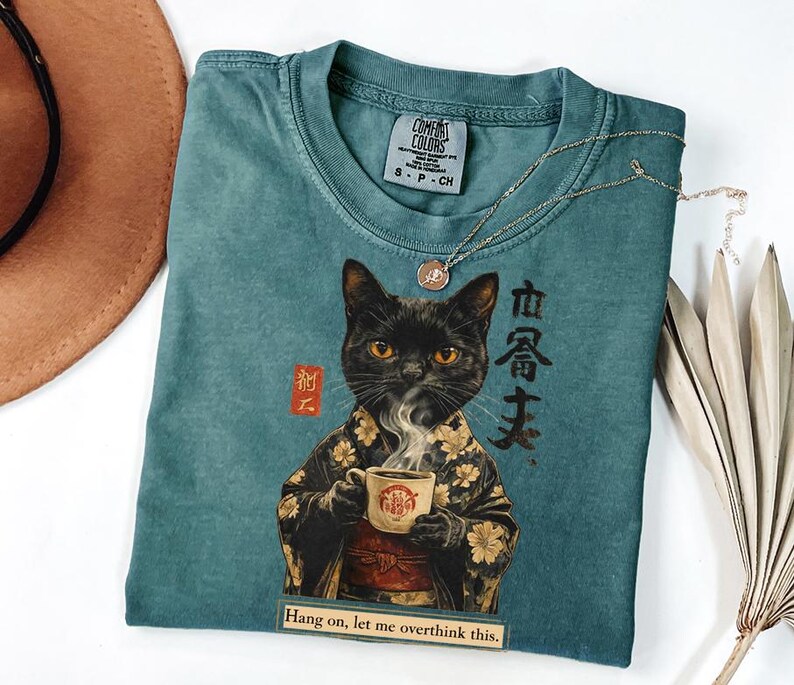 May include: Teal Comfort Colors t-shirt featuring a black cat in a kimono holding a coffee cup. The shirt has Japanese text and the phrase "Hang on, let me overthink this." A gold necklace and a hat are also in the image.