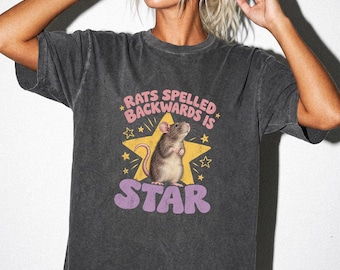 May include: A charcoal gray short-sleeve t-shirt featuring a graphic of a brown rat standing in front of a yellow star. The text reads RATS SPELLED BACKWARDS IS STAR in pink and purple lettering, surrounded by small yellow star icons.