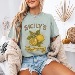 May include: A light green t-shirt with the text "SICILY'S Finest Lemons" and a graphic of lemons and leaves. The shirt is worn with denim shorts and a cream-colored cardigan. The overall theme is a casual, summery style.