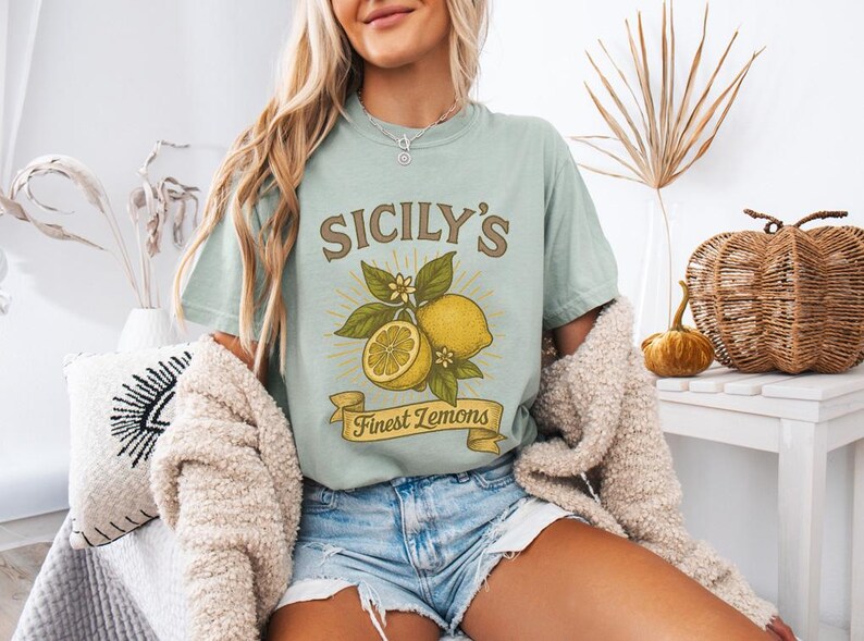 May include: A light green t-shirt with the text "SICILY'S Finest Lemons" and a graphic of lemons and leaves. The shirt is worn with denim shorts and a cream-colored cardigan. The overall theme is a casual, summery style.
