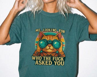 May include: A dark teal short-sleeve t-shirt featuring a graphic of an orange cat looking through blue binoculars. The text reads ME, LOOKING FOR WHO THE FUCK ASKED YOU. The design includes a circular sunset background with orange, pink, and teal.