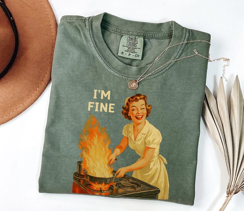 May include: Sage green t-shirt with the phrase "I'M FINE" and a vintage illustration of a woman cooking with flames. A silver necklace with a pendant is on the shirt. A brown hat is in the upper left corner.