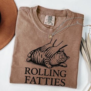 May include: A brown Comfort Colors t-shirt with a graphic of a cat being petted. The cat is lying down and smiling. The text "ROLLING FATTIES" is printed below the cat in a bold font. A silver necklace with a small pendant is on the shirt.