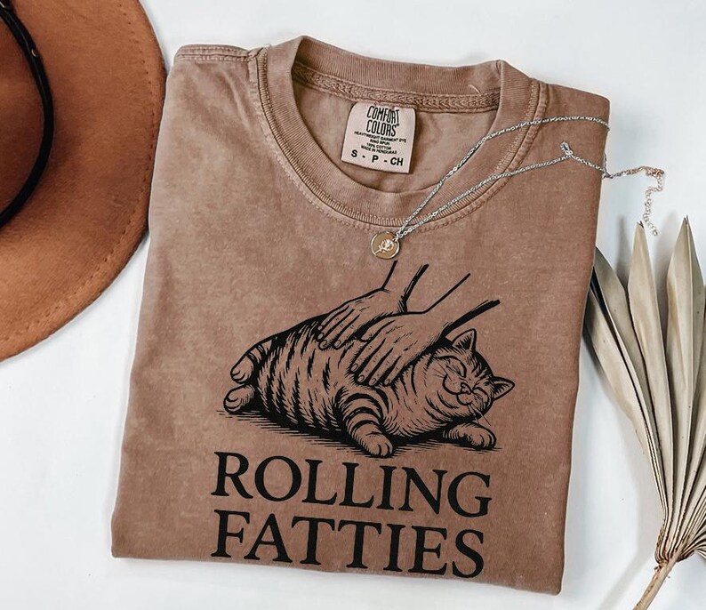 May include: A brown Comfort Colors t-shirt with a graphic of a cat being petted. The cat is lying down and smiling. The text "ROLLING FATTIES" is printed below the cat in a bold font. A silver necklace with a small pendant is on the shirt.