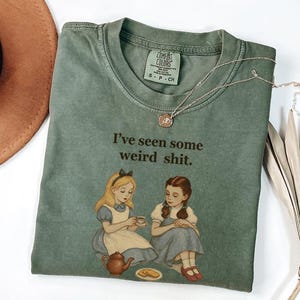 May include: An olive green t-shirt with the text "I've seen some weird shit" and a graphic of two girls. The shirt has a crew neck and short sleeves. A gold necklace is draped across the shirt.