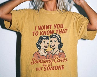 May include: A mustard yellow short-sleeve t-shirt featuring a graphic of two women with retro hairstyles. The text reads: I WANT YOU TO KNOW THAT Someone Cares NOT ME BUT SOMONE. The shirt has a crew neckline and a relaxed, loose-fitting silhouette.