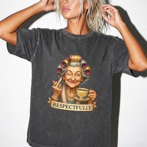 Puede incluir: Una camiseta gris carbón de manga corta con el gráfico de una persona mayor con rulos, sosteniendo una taza de té, guiñando un ojo y haciendo un gesto con el dedo medio. Debajo del gráfico hay una cinta con el texto "RESPECTFULLY".