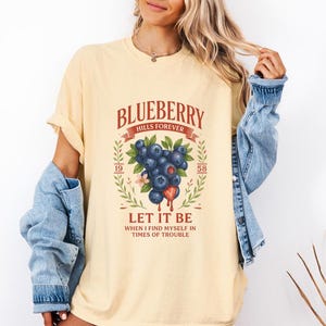 May include: Cream-colored t-shirt featuring a graphic design with the text "BLUEBERRY HILLS FOREVER" above a cluster of blueberries and the words "LET IT BE" below. The design includes the numbers "19" and "58".