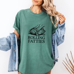 May include: A teal-colored t-shirt featuring a black illustration of a cat being massaged, with the text "ROLLING FATTIES" below. The shirt is worn by a person with blonde hair and a denim jacket.