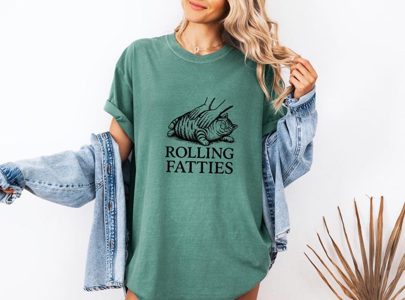 May include: A teal-colored t-shirt featuring a black illustration of a cat being massaged, with the text "ROLLING FATTIES" below. The shirt is worn by a person with blonde hair and a denim jacket.