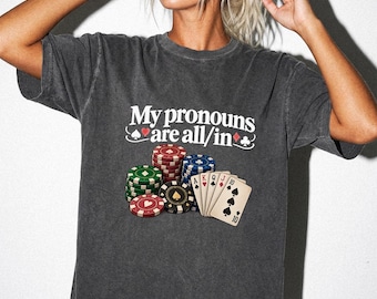 May include: A charcoal gray short-sleeve t-shirt featuring the white text My pronouns are all/in with red and black card suit symbols. Below the text is a graphic of stacked red, green, blue, and black poker chips and five playing cards.