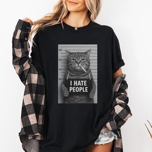May include: Black t-shirt featuring a black and white image of a cat in a mugshot, holding a sign that reads "I HATE PEOPLE." The cat is smoking a cigarette. The shirt is worn by a person wearing a black and white plaid shirt.