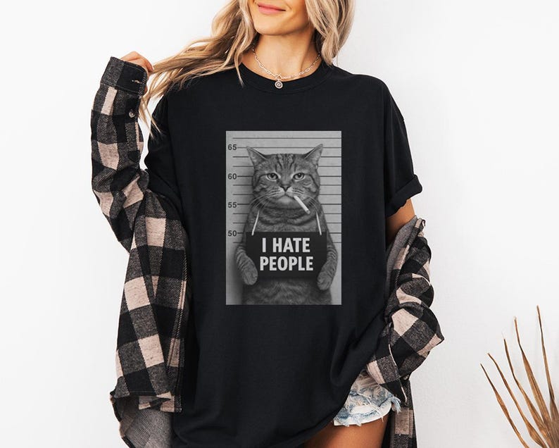 May include: Black t-shirt featuring a black and white image of a cat in a mugshot, holding a sign that reads "I HATE PEOPLE." The cat is smoking a cigarette. The shirt is worn by a person wearing a black and white plaid shirt.