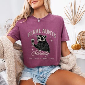 May include: A mauve-colored t-shirt with the words "FERAL AUNTS Society" and a graphic of a raccoon holding a wine glass. The raccoon is black and white. The text "EXCLUSIVE MEMBERS ONLY" is below the raccoon.