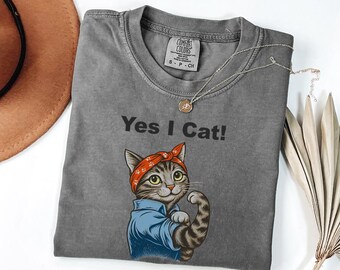 May include: A gray t-shirt with the text "Yes I Cat!" and a cartoon cat flexing its arm. The cat wears a red bandana and a blue shirt. A gold necklace is draped across the shirt.