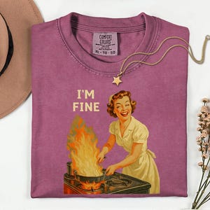May include: A mauve Comfort Colors t-shirt with the text "I'M FINE" and a vintage illustration of a woman cooking with flames. A gold star pendant necklace is draped across the shirt.