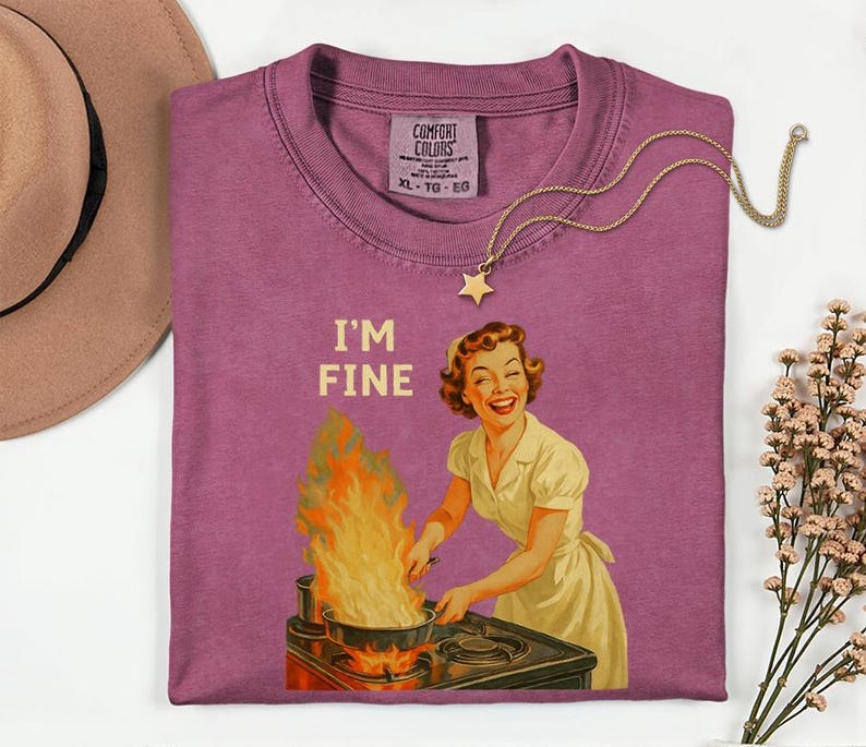 May include: A mauve Comfort Colors t-shirt with the text "I'M FINE" and a vintage illustration of a woman cooking with flames. A gold star pendant necklace is draped across the shirt.