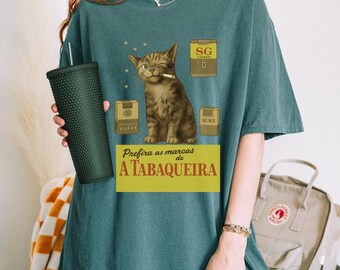 Op de afbeelding: Tealkleurig T-shirt met een kat die een sigaret rookt, omringd door vintage sigarettendozen. De tekst "Prefira as marcas de A TABAQUEIRA" staat op een gele banner. Een donkergroene beker wordt in de linkerhand gehouden.