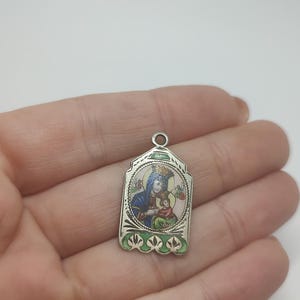 May include: A silver pendant featuring a religious image of a woman and child, with a blue and red color scheme. The pendant has a rectangular shape with a decorative border and a small loop at the top for a chain.