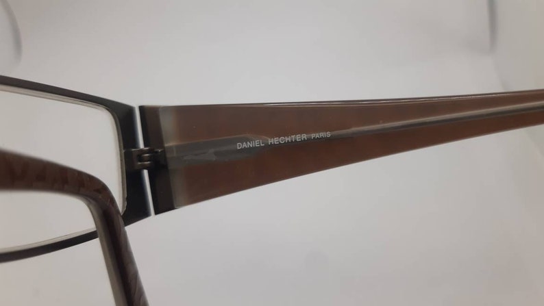 May include: Brown and black eyeglasses with a brown plastic frame and metal arms. The frame has the text "DANIEL HECHTER PARIS" printed on it.
