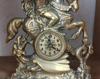 Könnte beinhalten: Eine Messinguhr mit einem Ritter, der einen Drachen erschlägt. Das Zifferblatt der Uhr hat schwarze römische Ziffern und ein Schneeflockenmuster in der Mitte.
