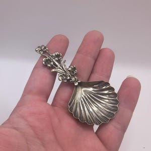 May include: A silver-toned metal spoon featuring a bowl shaped like a scalloped seashell. The handle of the spoon is decorated with a detailed floral motif consisting of several small flowers and leaves extending from the base of the bowl.