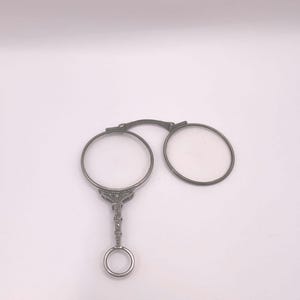 Könnte beinhalten: Eine Lorgnette aus silberfarbenem Metall mit zwei kreisförmigen Rahmen, die durch einen Steg verbunden sind, sowie einem dekorativen, strukturierten Griff, der nach unten verläuft und in einem kleinen Ring zum Halten endet.