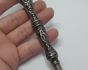 May include: Ornate silver-toned tool with a pointed end and detailed floral and scrollwork patterns. The handle has a cylindrical shape with decorative bands. The tip has a small opening. The tool is likely made of sterling silver, as indicated by the "925" marking.